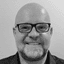 Bald man with glasses and a beard smiling at the camera in black and white.